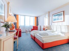 Einblick in das lichtdurchflutete Zimmer mit Doppelbett unter einem Gemälde gegenüber eines Schreibtischs mit Fernseher