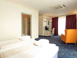 Doppelbett gegenüber eines orangefarbenen Sessels an einem Fenster mit roten Vorhängen 